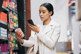 Mujer en un supermercado comparando precios en su teléfono, destacando el aumento del coste de los alimentos.